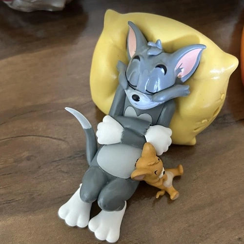 Close-up top view of the Tom and Jerry dashboard toy PVC figurine, showing the detailed sleeping pose of Tom cat reclined on a yellow pillow with Jerry mouse resting beside his leg, placed on a wood grain table surface.