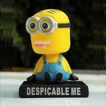 A cute Minion auto interior decoration nodding figure sitting on a dashboard surface with a blurred outdoor background.