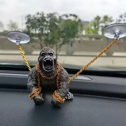 A silver ape dashboard ornament securely positioned inside a car, featuring the roaring figure restrained by gold chains attached to the windshield via suction cups.